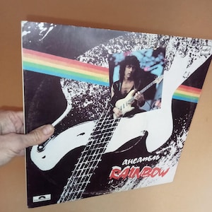 Könnte beinhalten: Ein schwarzes und weißes Vinyl-Plattencover mit einem Regenbogenstreifen-Design. Das Cover zeigt ein Foto eines Mannes, der eine weiße E-Gitarre spielt. Der Text "ансамбль RAINBOW" ist auf dem Cover gedruckt.