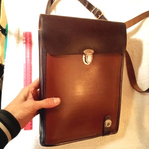 May include: A brown leather messenger bag with a dark brown flap and a shoulder strap. The bag has a metal clasp closure and a small button closure on the bottom. The bag is approximately 12 inches tall and 10 inches wide.