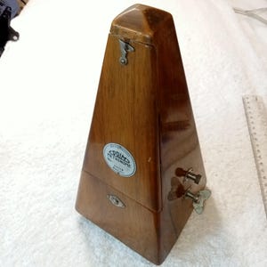Può includere: Un metronomo vintage in legno a forma di piramide. Il metronomo è realizzato in legno lucido con venature visibili. Ha un fermo in metallo e un'etichetta che recita "Codim Metronome". Due manopole in metallo sono visibili sul lato.