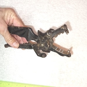 May include: A vintage, black dragon-shaped nutcracker. The nutcracker features a detailed dragon head with open jaws and a textured body. The handle has a wing-like design. The metal tool is held in a hand.