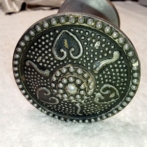 May include: Close-up of a silver and black metal bottle stopper with a decorative design. The stopper has a circular base with a raised center and a ring of small dots around the edge.