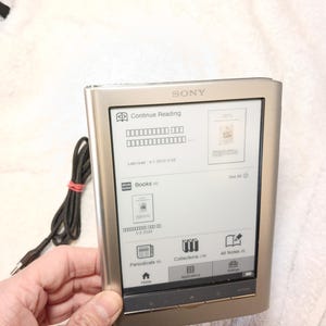 Puede incluir: Lector de libros electrónicos Sony plateado con pantalla negra que muestra una interfaz de libro. El dispositivo tiene un marco metálico y botones de navegación en la parte inferior. Se ve un cable negro con una banda roja.