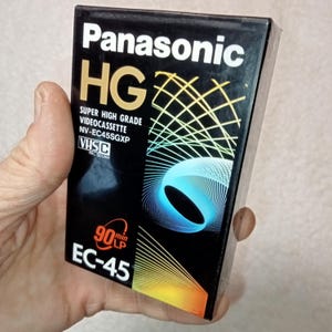Könnte beinhalten: Ein schwarzes VHS-Videokassettenband mit der Aufschrift "Panasonic HG" und einer Aufnahmezeit von 90 Minuten. Das Band ist mit "Super High Grade Video Cassette NV-EC45SGXP" beschriftet und kompatibel mit VHS-C- und PAL/SECAM-Systemen.