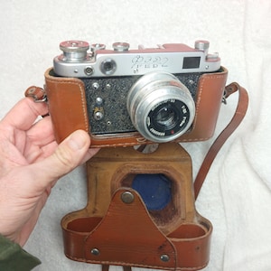 May include: A vintage FED 2 rangefinder camera with a silver top plate and black textured body, housed in a brown leather case with a matching shoulder strap. The lens is marked with the text Industar-61, 1:2.8, F=5cm, and serial number.
