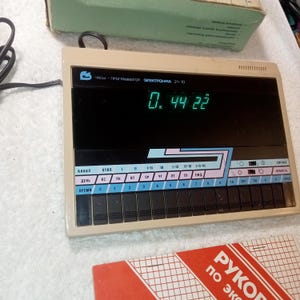 May include: A retro electronic clock and programmer, model 21-10, in a beige casing, displaying the time on a black screen. It has multiple buttons and Cyrillic text labels. The clock is partially visible with a box in the background.