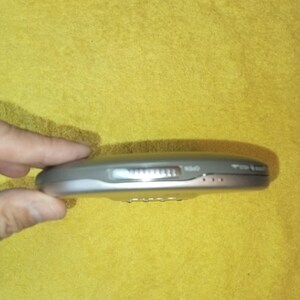 May include: A silver portable CD player with a volume knob and buttons for play, stop, and skip. The player is on a yellow surface.