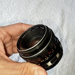 May include: A black Pentacon auto 1.8/50 camera lens with a silver ring and red markings. The lens has a depth of field scale and aperture markings.
