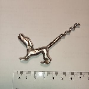 May include: A silver-colored dog-shaped corkscrew. The dog is in a playful pose, with its head up and tail extended. The corkscrew is attached to the dog's back. The ruler next to the corkscrew measures up to 8 cm.