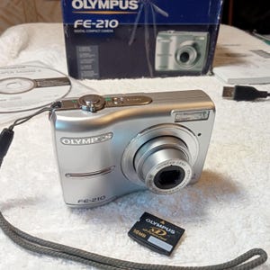Puede incluir: Cámara digital compacta plateada Olympus FE-210 con lente, flash y tarjeta XD-Picture de 16 MB. La cámara está sobre una superficie blanca, con la caja original y un CD en el fondo. La cámara tiene un objetivo zoom.
