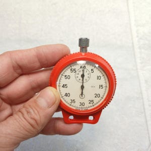 May include: A round mechanical stopwatch with a bright red plastic casing and a white face. The dial features black numerals from 5 to 60, a smaller sub-dial, and a silver metal winding crown at the top. The text MADE IN USSR is printed at the bottom.
