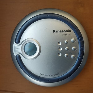 Puede incluir: Un reproductor de CD port&aacute;til plateado y azul con el texto "Panasonic SL-SX320" y "Anti-Skip System". El reproductor tiene varios botones, incluyendo reproducir, detener, saltar y repetir.
