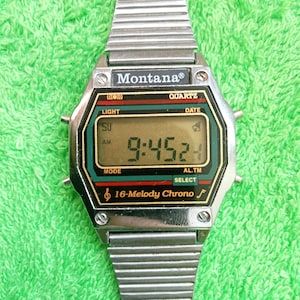 May include: A silver Montana digital watch with a rectangular face showing the time 9:45:24. The watch face is black, green, and red, with the text "Montana", "Quartz", and "16-Melody Chrono". The watch strap is made of silver metal.