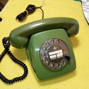 May include: Green vintage rotary dial telephone with a black coiled handset cord and a straight cord. The dial features numbers and the text "FEUER NOTRUF". The phone is on a yellow surface.
