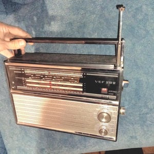Può includere: Una radio vintage argentata e nera con un quadrante cromato e un'antenna telescopica. La radio ha una faccia nera con il testo "VEF 202" stampato su di essa.