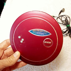 May include: A red portable CD player with the text "MP3 CD-DA VCD" on a blue sticker. The player has a silver trim and the brand name "BBK". Black earbuds are connected to the player.