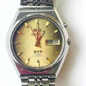 Puede incluir: Un reloj vintage Orient Crystal 21 Jewels con esfera dorada y correa de acero inoxidable. La esfera muestra el día y la fecha, con detalles en rojo en las agujas y marcadores. La caja del reloj es plateada.