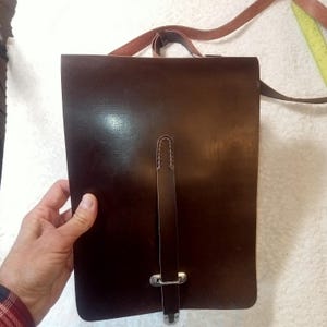 May include: A dark brown leather satchel with a long shoulder strap. The bag has a rectangular shape with a flap closure and a vertical strap secured by a metal clasp. The bag is held by a person's hand.