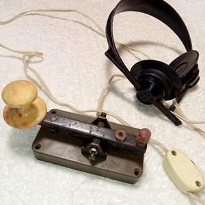 May include: Vintage telegraph key and headset. The key is a rectangular metal base with a lever and a marbled knob. The headset is black with ear cups and a headband. Both are connected by wires.