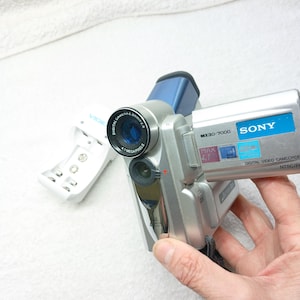 Könnte beinhalten: Silberner digitaler Sony-Videocamcorder mit blauem Objektiv und weißem Batterieladegerät. Auf dem Camcorder steht "MX30-7000" und "SONY". Das Objektiv trägt die Aufschrift "DIGITAL CAMERA 0,9 mm f/1,8 4,1 MEGAPIXEL".