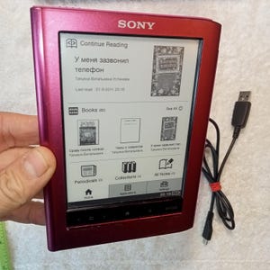 Puede incluir: Lector de libros electrónico Sony rojo con texto en ruso, incluyendo "У меня зазвонил телефон". La pantalla muestra portadas de libros y opciones de menú. Un cable USB está conectado.