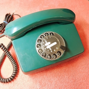 May include: A vintage, teal-colored rotary dial telephone. The phone features a handset, a base with a circular dial, and a coiled cord. The dial has black numbers and a clear plastic cover. The phone is on a red surface.