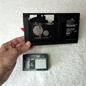 May include: A black Panasonic cassette adapter with the text "CASSETTE ADAPTOR" and a small JVC VHS-C video cassette. The adapter is open, revealing its interior. The JVC cassette is labeled "45 VHS-C".