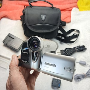 Op de afbeelding: Een zilverkleurige Panasonic NV-GS6 MiniDV-camcorder met een 20x optische zoomlens, een uitklapbaar LCD-scherm en een stereo zoommicrofoon. De set bevat een zwarte draagtas, een acculader, een accu en een zwarte stroomkabel.