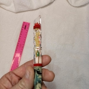 Puede incluir: Un bolígrafo decorativo transparente con un acento rojo y blanco y una base veteada azul y verde. Una regla rosa está en el fondo. El bolígrafo está en una mano.