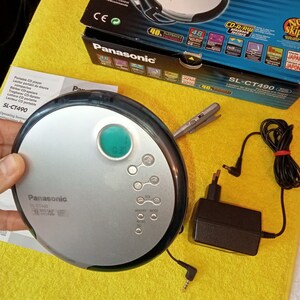 May include: A silver and black Panasonic SL-CT490 portable CD player with a green display screen and buttons for volume, memory, mode, and repeat. The player is plugged into a black power adapter with a cord.