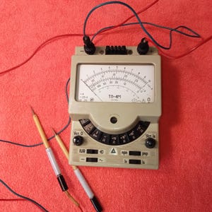 Puede incluir: Un multímetro analógico beige vintage con una esfera circular y cables negros y rojos. El medidor tiene una serie de configuraciones y una aguja que indica las mediciones. Dos sondas de prueba están colocadas cerca del medidor sobre una superficie roja.