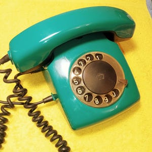 May include: A vintage teal rotary dial telephone with a black coiled cord. The phone features a circular dial with white numbers and a black center. The handset is curved and rests on the phone's body. The phone is on a yellow surface.