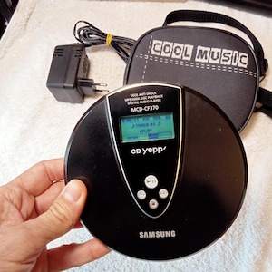 Può includere: Lettore CD Samsung nero con display digitale che mostra le informazioni sulla traccia. Il lettore è etichettato "CD yepp'" e "MCD-CF370". Include una custodia nera con "COOL MUSIC" stampato sopra e un adattatore di alimentazione nero.