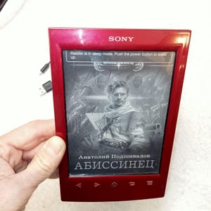 Puede incluir: Lector de libros electrónicos Sony rojo que muestra la portada de un libro con un hombre con una bufanda. La pantalla dice: "Reader is in sleep mode. Push the power button to wake up." Se ve un cable USB.