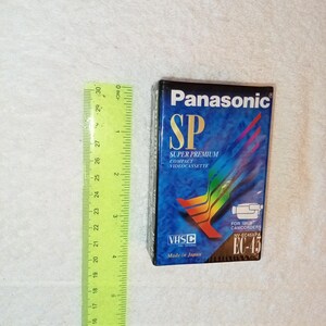 Könnte beinhalten: Eine Panasonic SP Super Premium Kompakt-Videokassette mit blauem und schwarzem Design. Das VHS-C-Band ist mit "Made in Japan" beschriftet und enthält Text für NV-EC45XPA Camcorder. Ein grünes Lineal liegt neben dem Band.