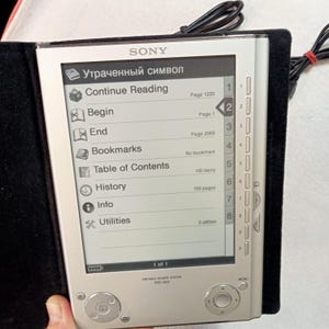 Puede incluir: Lector de libros electrónicos Sony PRS-505 plateado con una funda negra. La pantalla muestra un menú con opciones como "Continue Reading" y "Table of Contents". El dispositivo tiene botones físicos y un cable de alimentación.