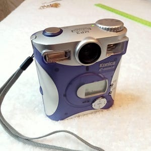 May include: A purple and silver Konica Minolta digital camera with a built-in MP3 player. The camera has a strap and a zoom lens.