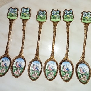 May include: Set of six gold-tone teaspoons with blue and pink bird designs on the bowls. Each spoon has a green and white enamel crest with a bird design on the handle.