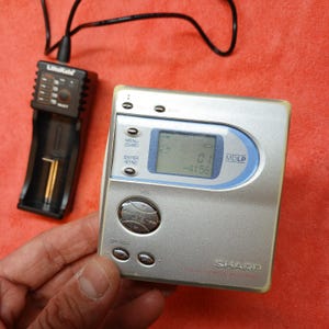 Könnte beinhalten: Ein silberner Sharp MDLP MiniDisc-Recorder mit einem blau akzentuierten Display, das Titelinformationen anzeigt. Ein schwarzes Ladegerät ist angeschlossen, und das Gerät hat mehrere Tasten und eine Lautstärkeregelung. Das Gerät ist vor rotem Hintergrund.