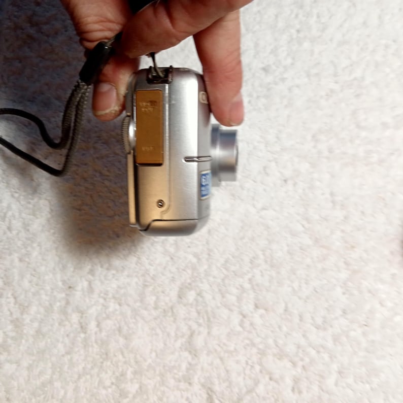 May include: A silver digital camera with a lens and a brown battery compartment. The camera is held by a black wrist strap. The camera has a small blue sticker on the side.