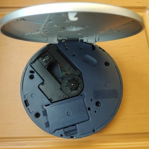 Puede incluir: Un reproductor de CD azul y plateado con la tapa abierta, que revela el mecanismo de la unidad de disco.