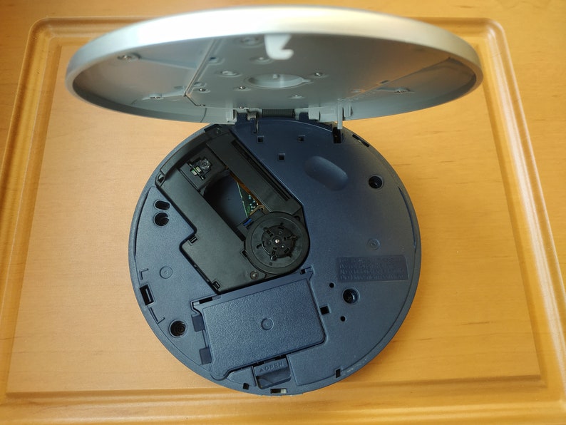 Puede incluir: Un reproductor de CD azul y plateado con la tapa abierta, que revela el mecanismo de la unidad de disco.