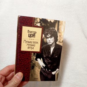 Peut inclure: Un livre intitulé "Viktor Tsoi" avec le texte "Muzyka voln, muzyka vetra" sur la couverture. La couverture présente un portrait d'un homme en chemise noire. Le livre a un dos bordeaux et une image sépia.
