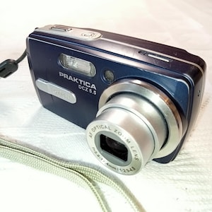 May include: A blue digital camera with a silver lens. The camera has the text "Praktica DCZ 5.5" on the front. The lens has the text "AF Optical Zoom Lens f=5.8-17.4mm 1:2.9-4.8" on it.