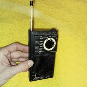 May include: A black and silver portable radio with a telescopic antenna. The radio has a dial with numbers from 200 to 550 and the text "ОЛИМПИК 402 КВ НАСТР ГРОМК" printed on the front.