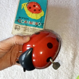 May include: A red and black wind-up toy ladybug with black spots and a key on the bottom. The toy is in its original box, which is blue and yellow and has the text "Жучок "СОЛНЫШКО"  КИЇВ  НИЕВСКИЙ МЕХАНИЧЕСКИЙ ЗАВОД ИГРУШЕК ИМ. Н.Ф. ВАТУТИН  ДЛЯ ДЕТЕЙ ОТ" printed on it.