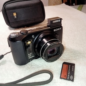 Puede incluir: Cámara digital Sony Cyber-shot negra con lente, flash y estuche de transporte negro. Una tarjeta Memory Stick PRO-HG Duo está al lado de la cámara. La cámara tiene impreso "Full HD Movie" y "Cyber-shot".