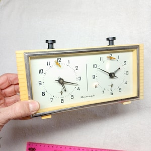 May include: A vintage chess clock with a cream-coloured frame and two clock faces. The clock faces have black numerals and hands, with a small gold-coloured accent at the top. The clock is rectangular and has two black buttons on top.