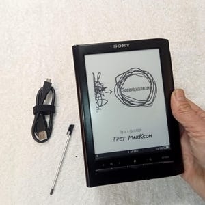 Puede incluir: Lector de libros electrónicos Sony negro con un lápiz óptico y cable de carga. La pantalla muestra la palabra "Эссенциализм" dentro de un círculo, con el nombre del autor, Greg McKeown, debajo. El lector es sujetado por una mano.