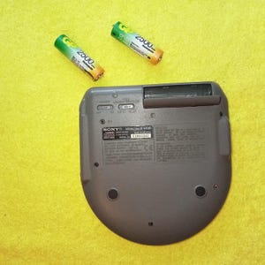 May include: A gray Sony CD player with the model number "D-E808" and serial number "15082397" printed on the back. The player has a slot for a CD and a compartment for two AA batteries.