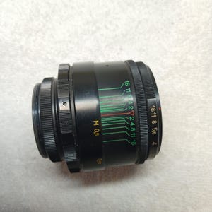 Peut inclure: Un objectif d'appareil photo noir avec des marquages verts et rouges et des chiffres blancs. L'objectif a une bague de mise au point stri&eacute;e et une bague d'ouverture plus petite. L'objectif porte les inscriptions "M 0,5" et "0,5".